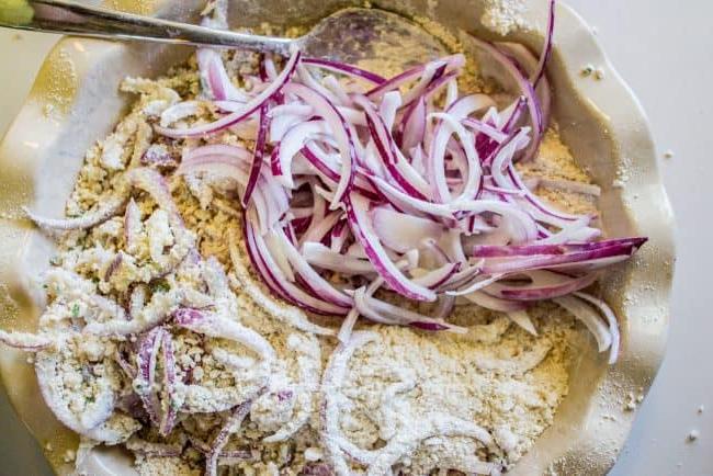 Επικάλυψη Κρεμμυδιών με Αλεύρι και Panko – Το μυστικό για τα τέλεια τραγανά κρεμμύδια. Προετοιμασία φετών κόκκινου κρεμμυδιού σε πιάτο πίτας με μεταλλική κουτάλα για τηγάνισμα.