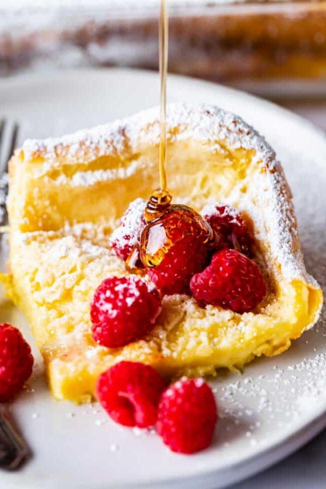 Dutch Baby τηγανίτα σε πιάτο με σμέουρα και ζάχαρη άχνη, περιχυμένη με σιρόπι.
