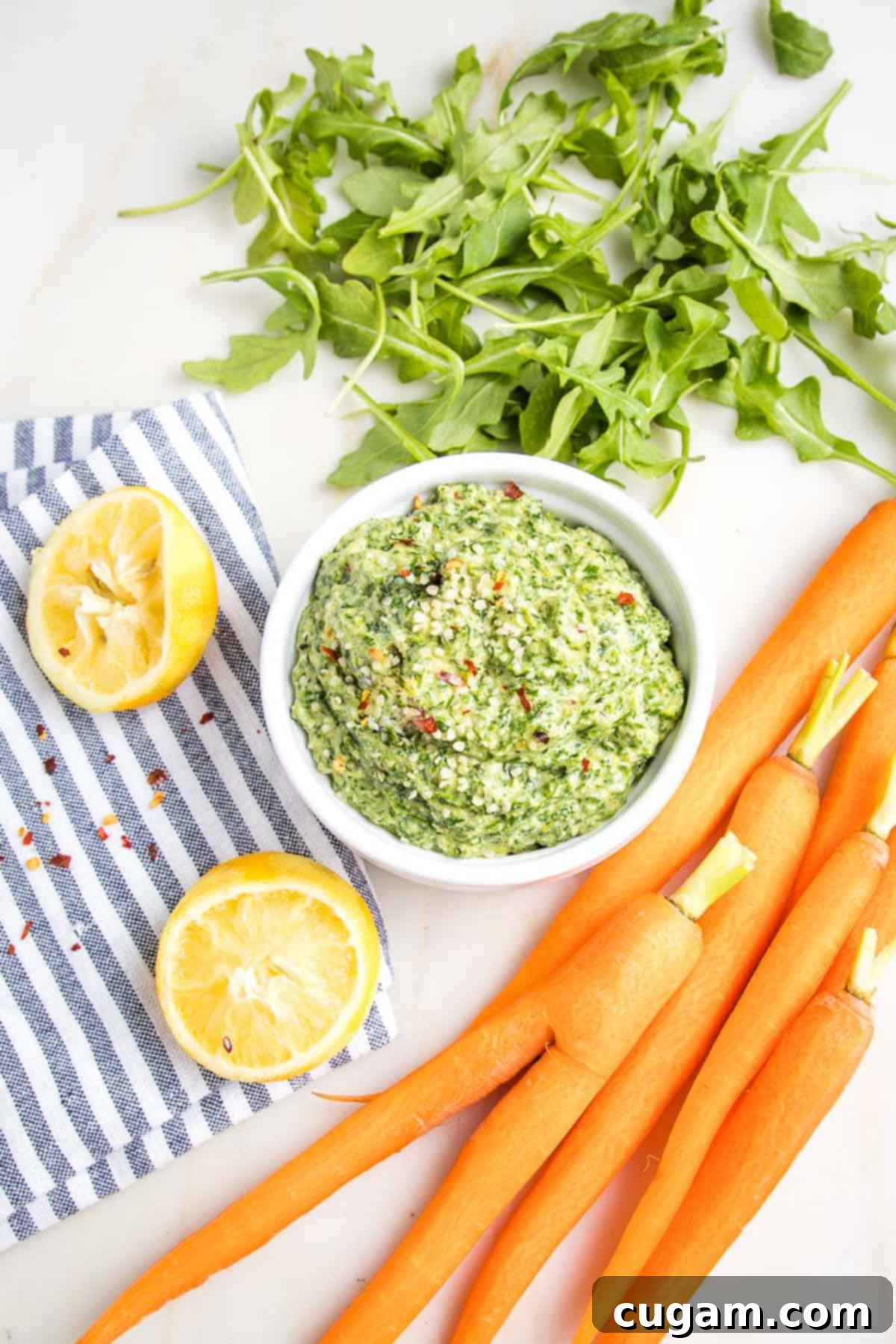 Freshly made carrot top pesto in a bowl, surrounded by carrots, carrot greens, and lemons, highlighting the main ingredients.