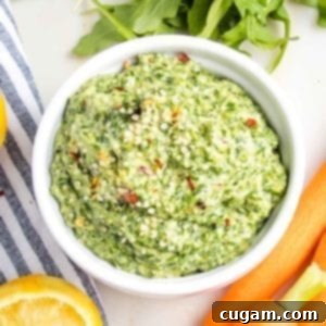 Close up of carrot top pesto in a small white bowl, sprinkled with hemp seeds and crushed red pepper. You can see glimpses of carrots, arugula and lemons surrounding the bowl.