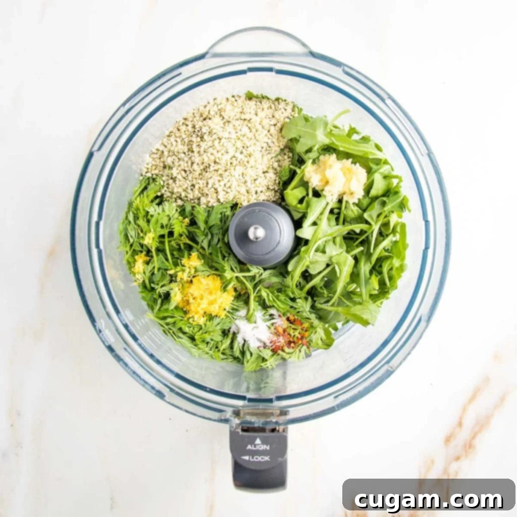 A food processor bowl filled with all the vibrant green ingredients for carrot greens pesto before blending: carrot tops, arugula, hemp seeds, lemon, and garlic.