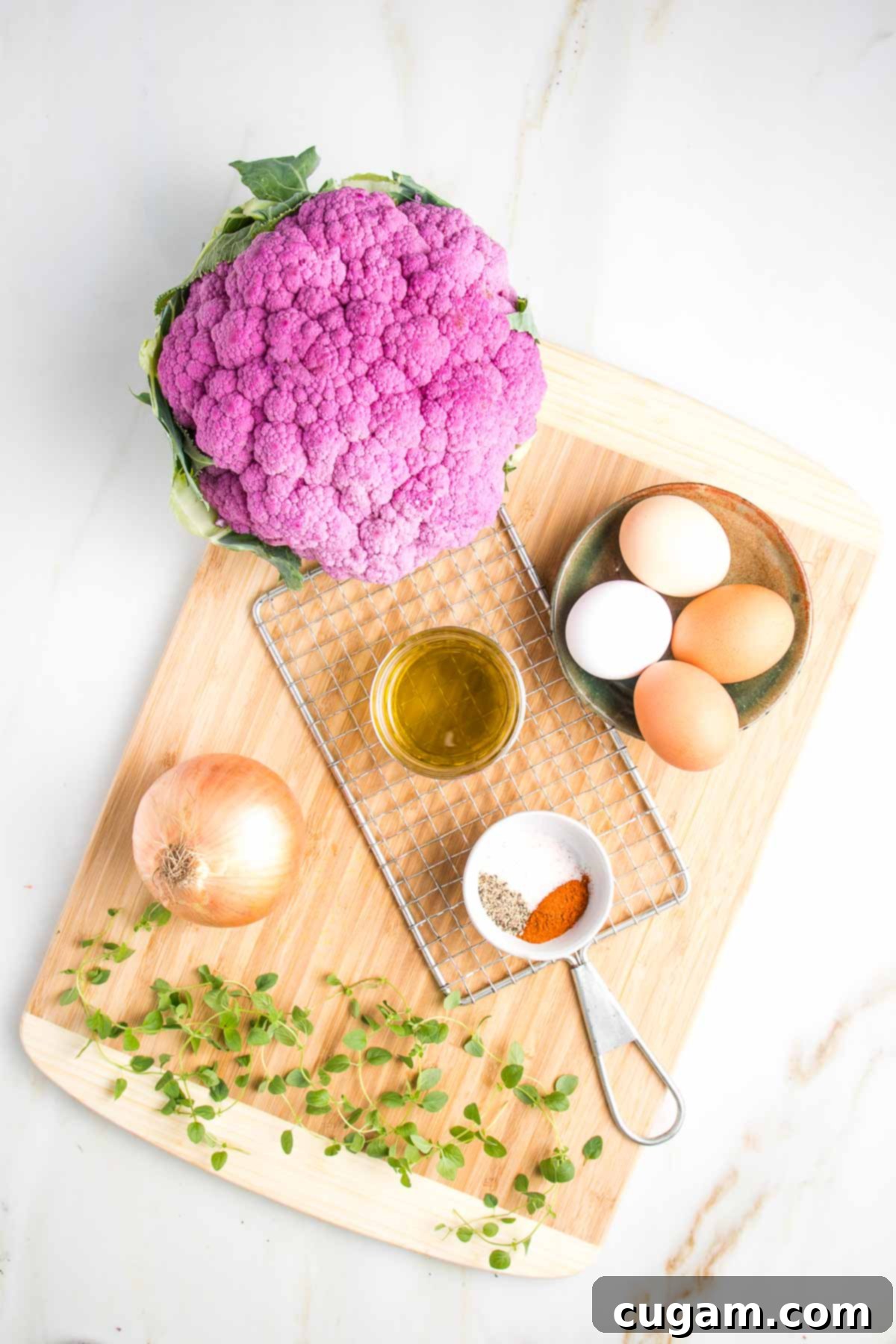Cauliflower kugel recipe ingredients on a cutting board: large head purple cauliflower, small ceramic dish with 4 eggs of varying colors from white to brown, sprigs of fresh oregano, small white dish with spices, small glass bowl of olive oil, a yellow onion and a potato shredder.