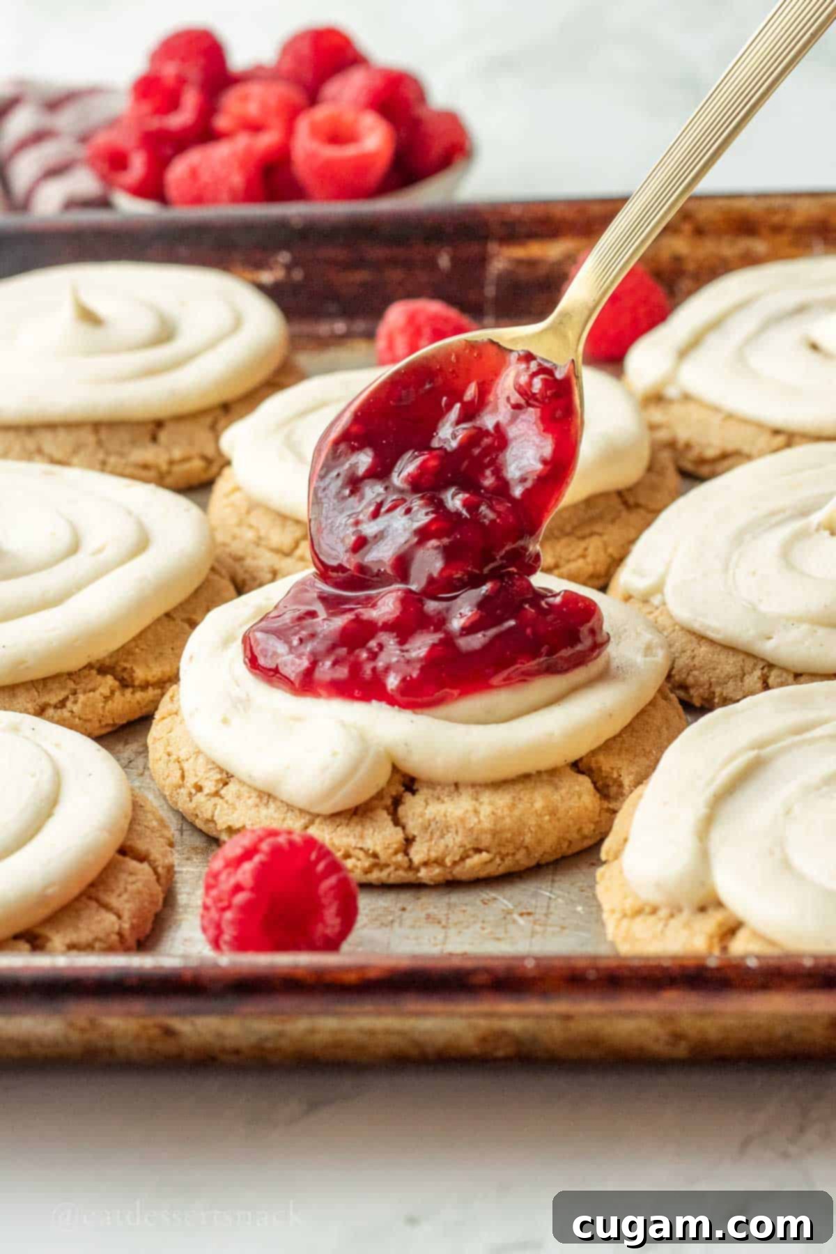 Creamy Raspberry Cheesecake Delights 9 A spoonful of vibrant red raspberry pie filling being carefully placed onto a frosted cheesecake cookie.