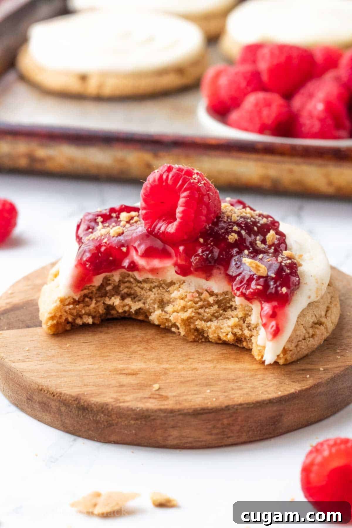 Creamy Raspberry Cheesecake Delights 10 A raspberry cheesecake cookie with a bite taken out, showcasing the distinct layers of graham cracker, cream cheese frosting, and raspberry filling, placed on a wooden cutting board.