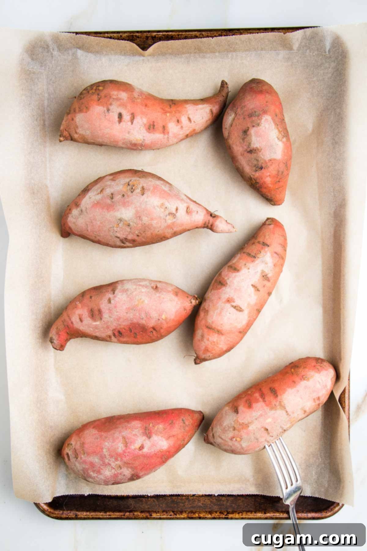 Whole sweet potatoes pricked with a fork on a parchment lined baking sheet.