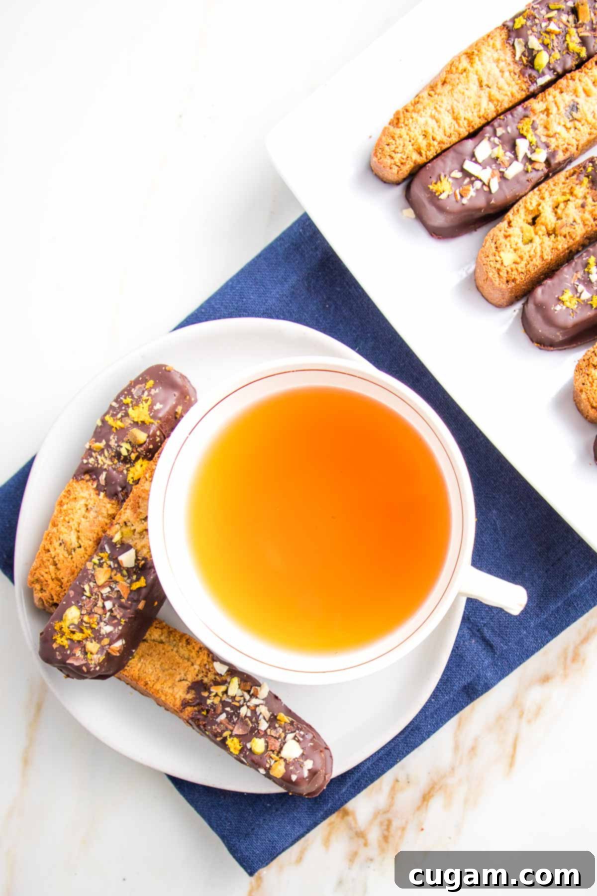 Biscotti, half dipped in chocolate and then sprinkled with chopped nuts and orange rind.  3 on a white plate with a mug of tea on the side.  Edge of platter showing with more of same biscotti.  Plates are resting on a dark blue cloth napkin. 