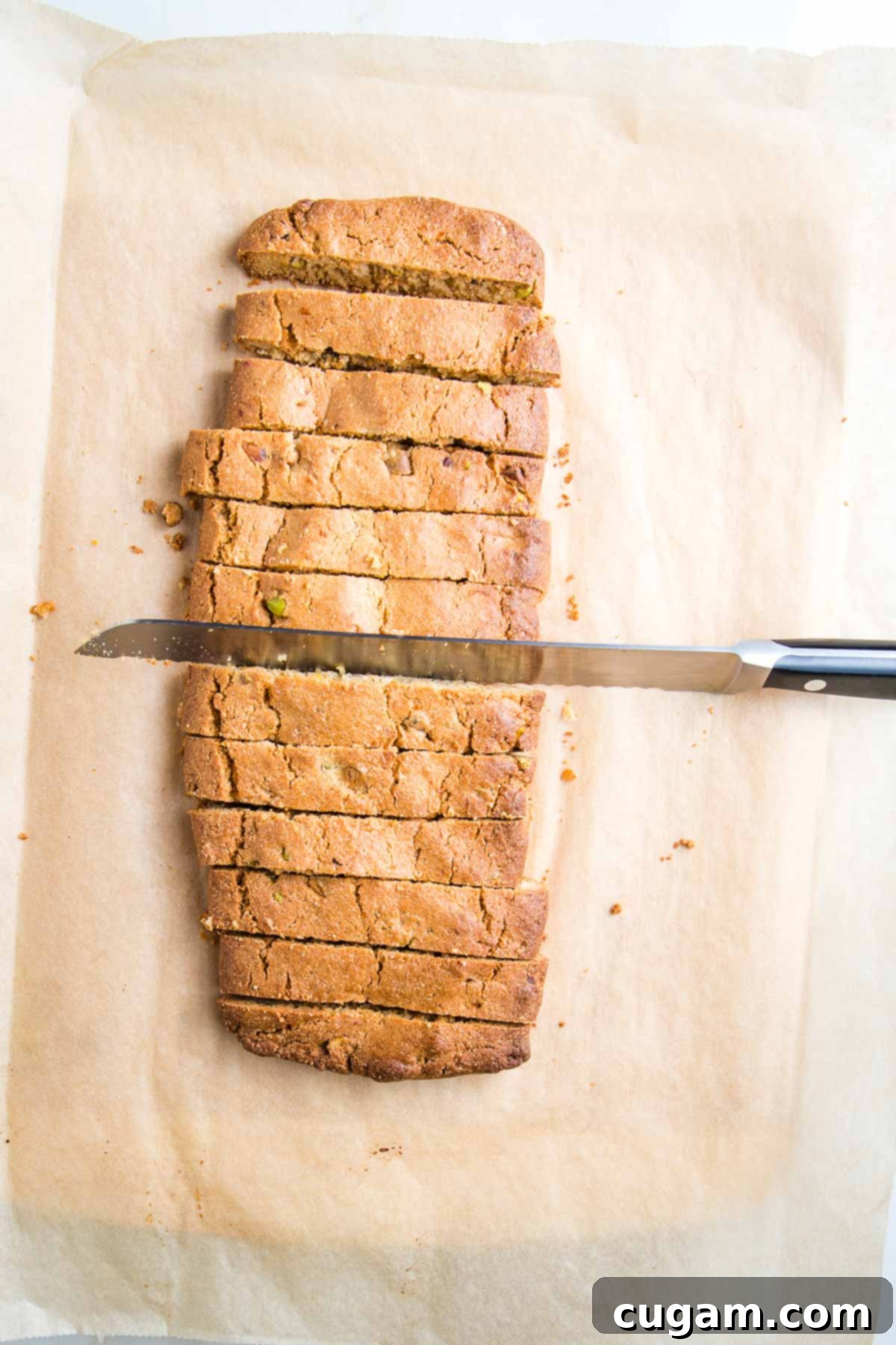 Log of first baked biscotti on a pieces of parchment, being sliced with a long serrated knife.