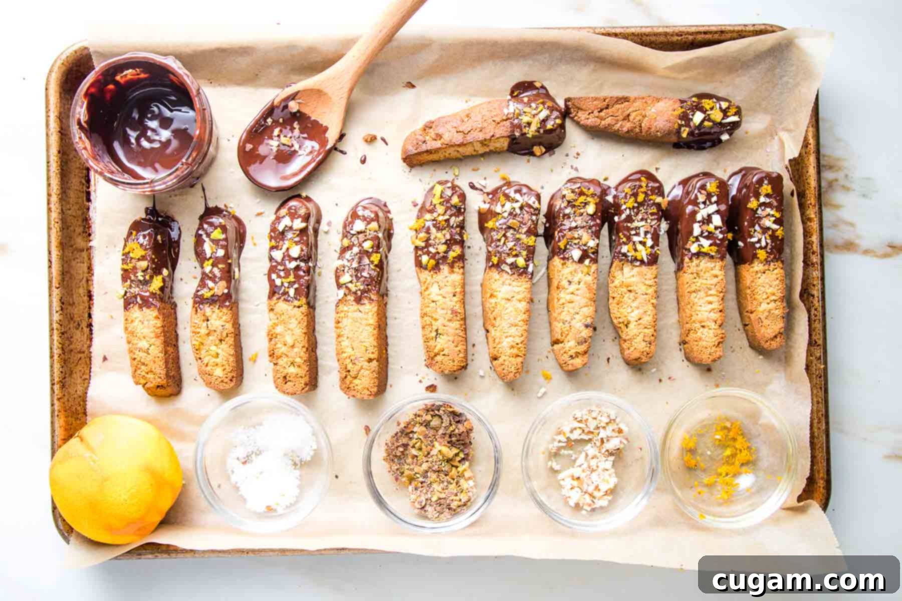 baking tray with biscotti cookies, small glass dish with melted chocolate and tiny bowls with orange zest, salt, chopped almonds and chopped pecans.  The biscotti have been dipped in chocolate and sprinkled with the toppings. 