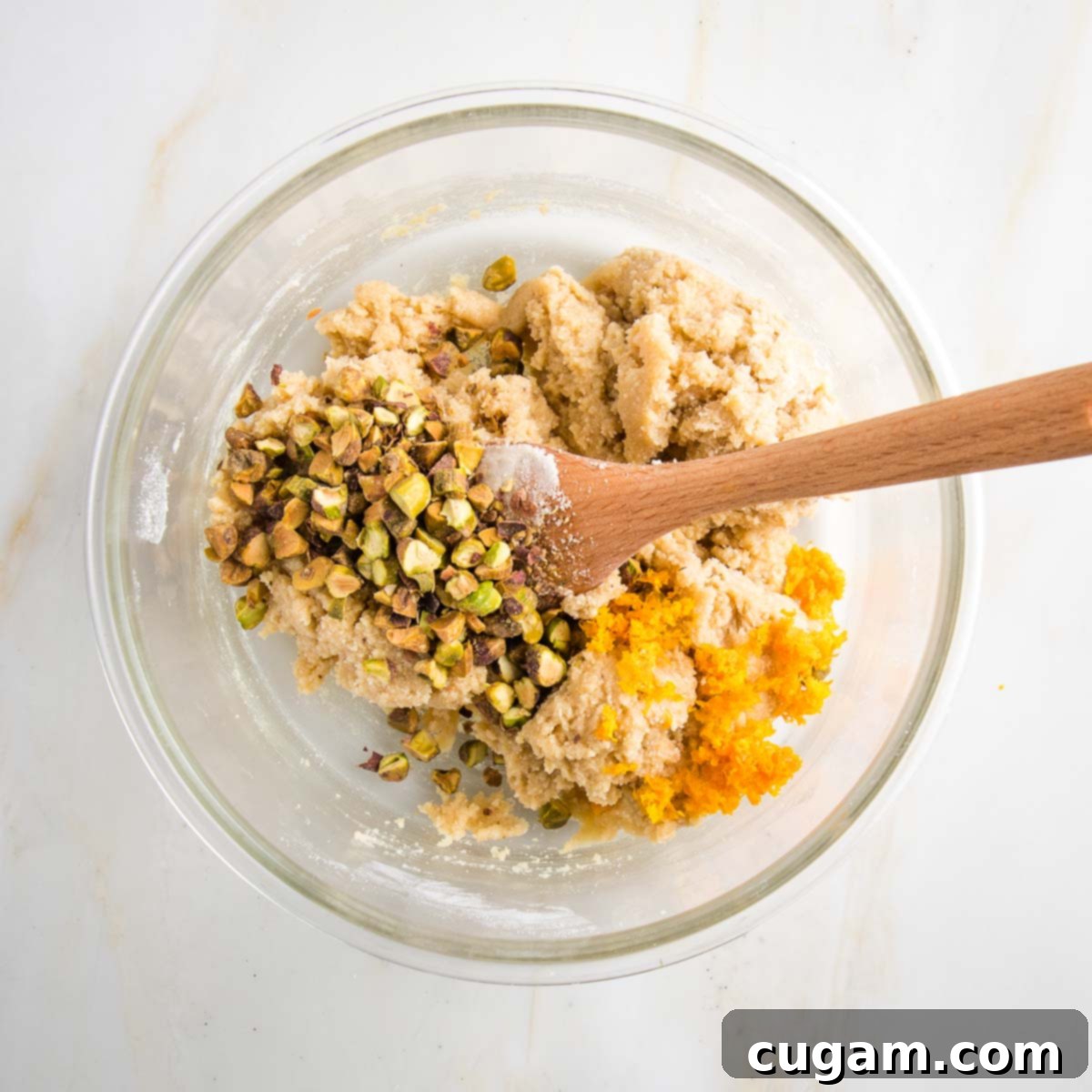 Gluten-free biscotti dough in a large glass bowl with added pistachios and orange zest.