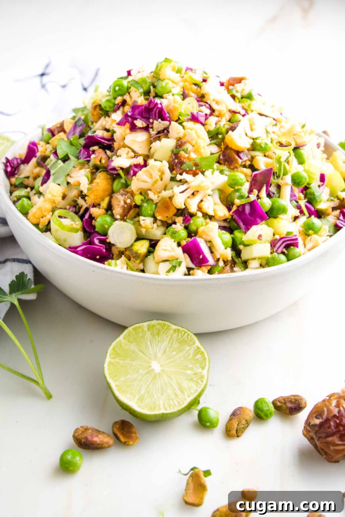 White bowl filled with colorful chopped salad that includes purple cabbage, fresh parsley, green peas, yellow cauliflower, pistachios, chopped dates, and scallions. There is a fresh slice of lime, some pistachios, dates and fresh parsley in the foreground on the white countertop