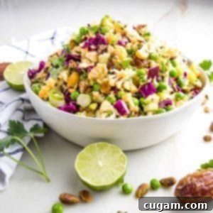 Side view of a white bowl that is filled with a colorful chopped salad that includes purple cabbage, yellow cauliflower, pistachios, fresh parsley, sliced scallions, green peas. Strewn around on the counter are fresh lime slices, dates, fresh parsley and a dish towel.