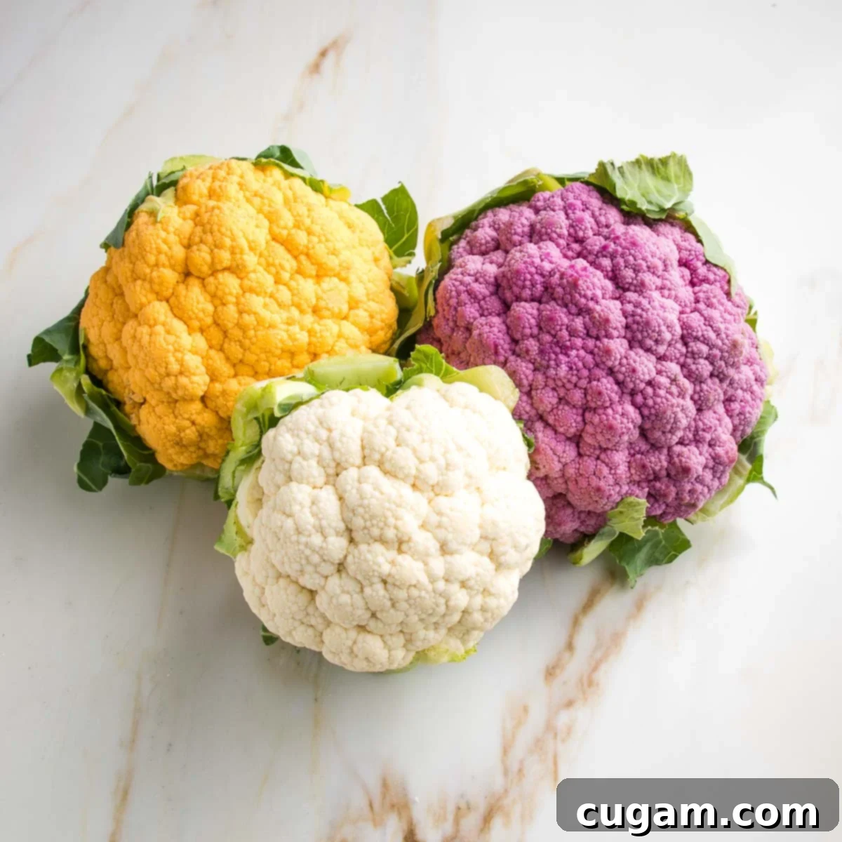 Three heads of cauliflower: purple, white, and yellow. All have their green leaves attached.