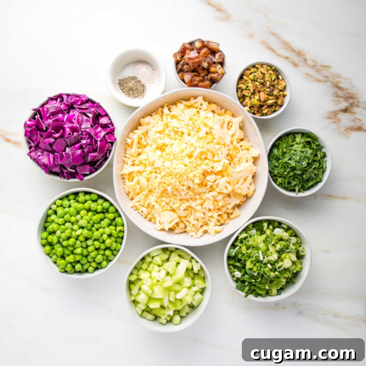 Bowls of ingredients for raw cauliflower salad. chopped cabbage, green peas, diced celery, sliced scallions, chopped parsley, pistachios, diced dates, salt and pepper. All small bowls positioned in a circle, surrounding a white bowl of shaved cauliflower.