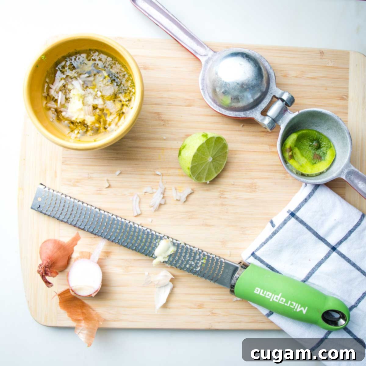 Small yellow bowl with lime vinaigrette. Also on the cutting board, are the ends of a shallot, a microplane zester with an end of a garlic clove and a juice squeezer with a squished lime inside. Plus a dish towel.
