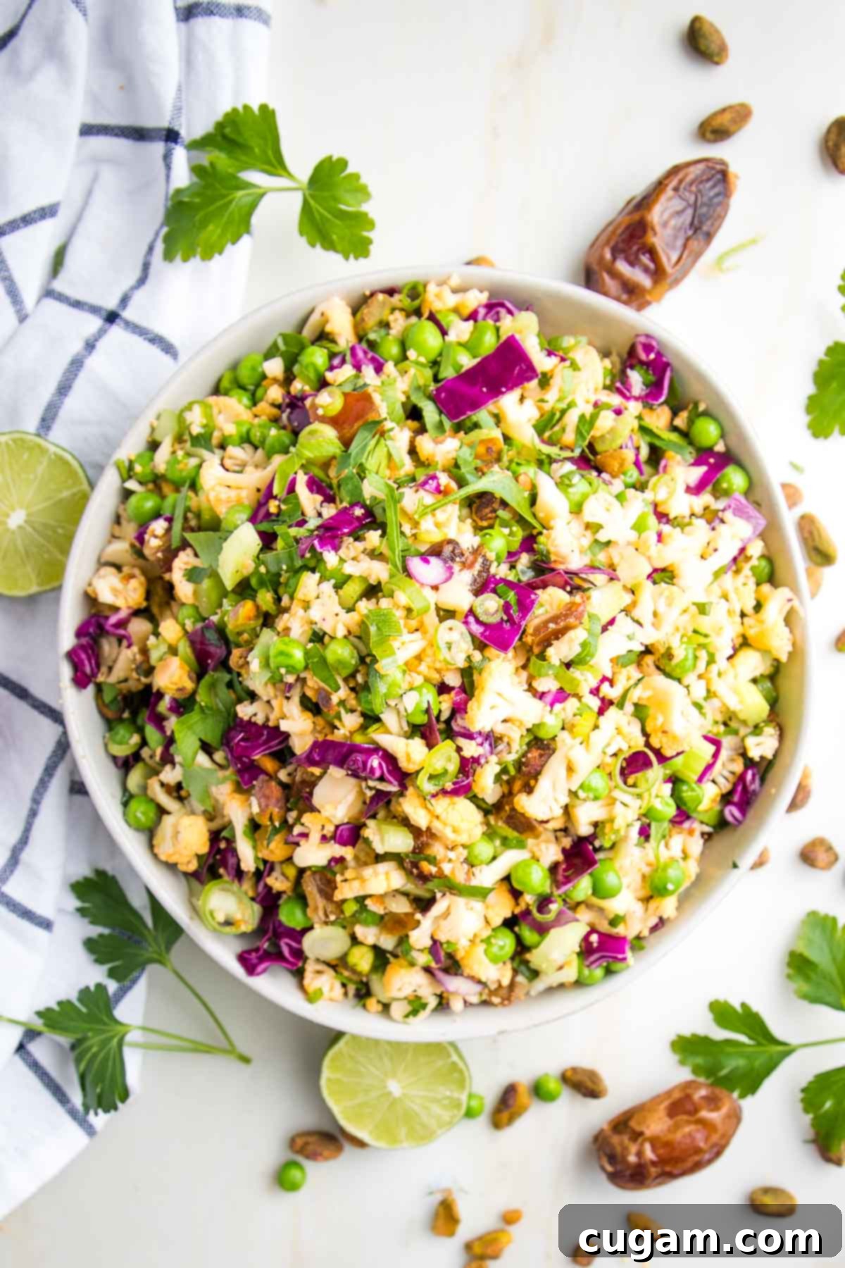 White bowl filled with raw cauliflower salad, including colorful veggies like purple cabbage, extra fresh cilantro, dates and pistachios along with a blue plaid dish towel surround the bowl.