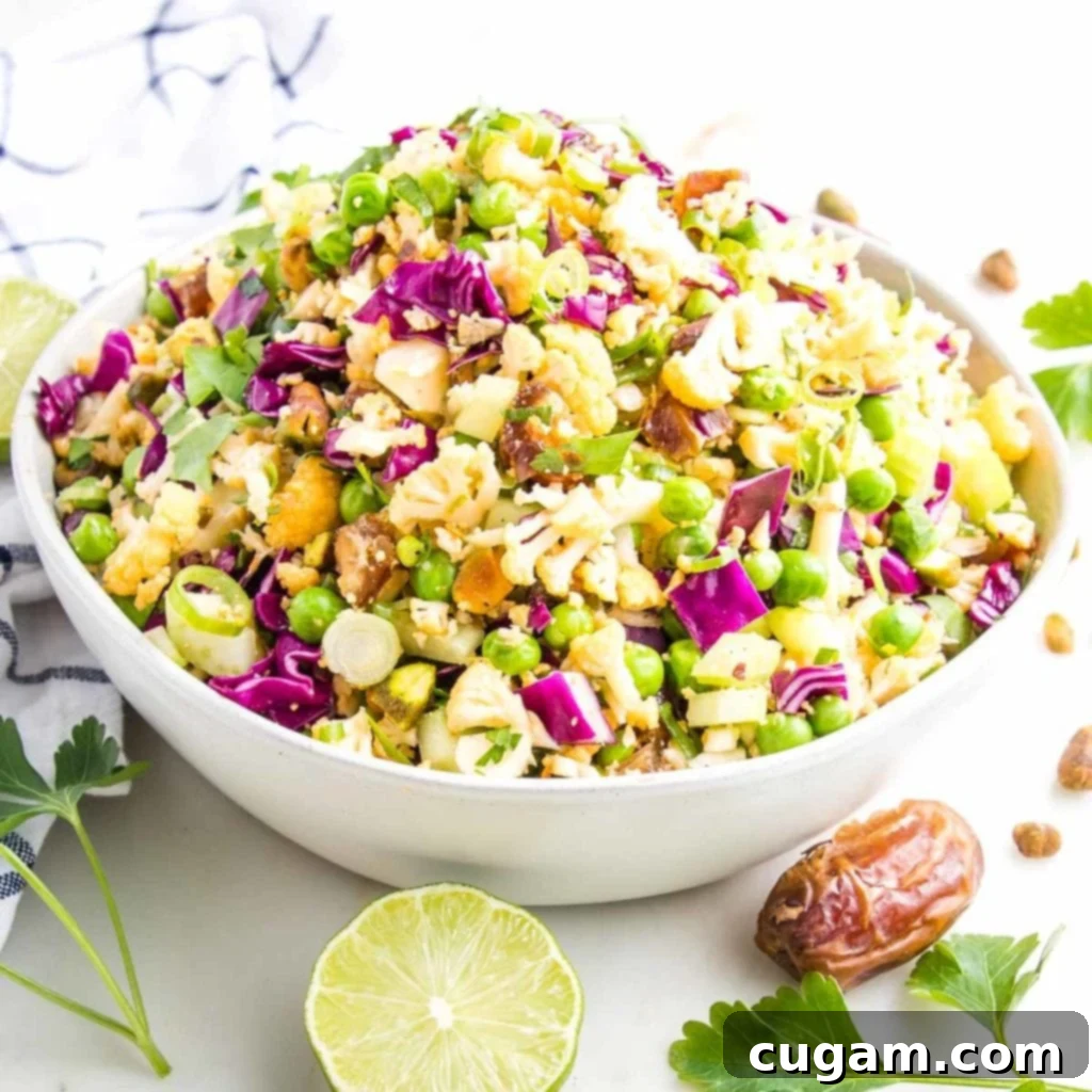 Large white bowl filled with colorful raw chopped cauliflower salad including cabbage, fresh herbs, scallions, green peas, pistachios and diced dates.
