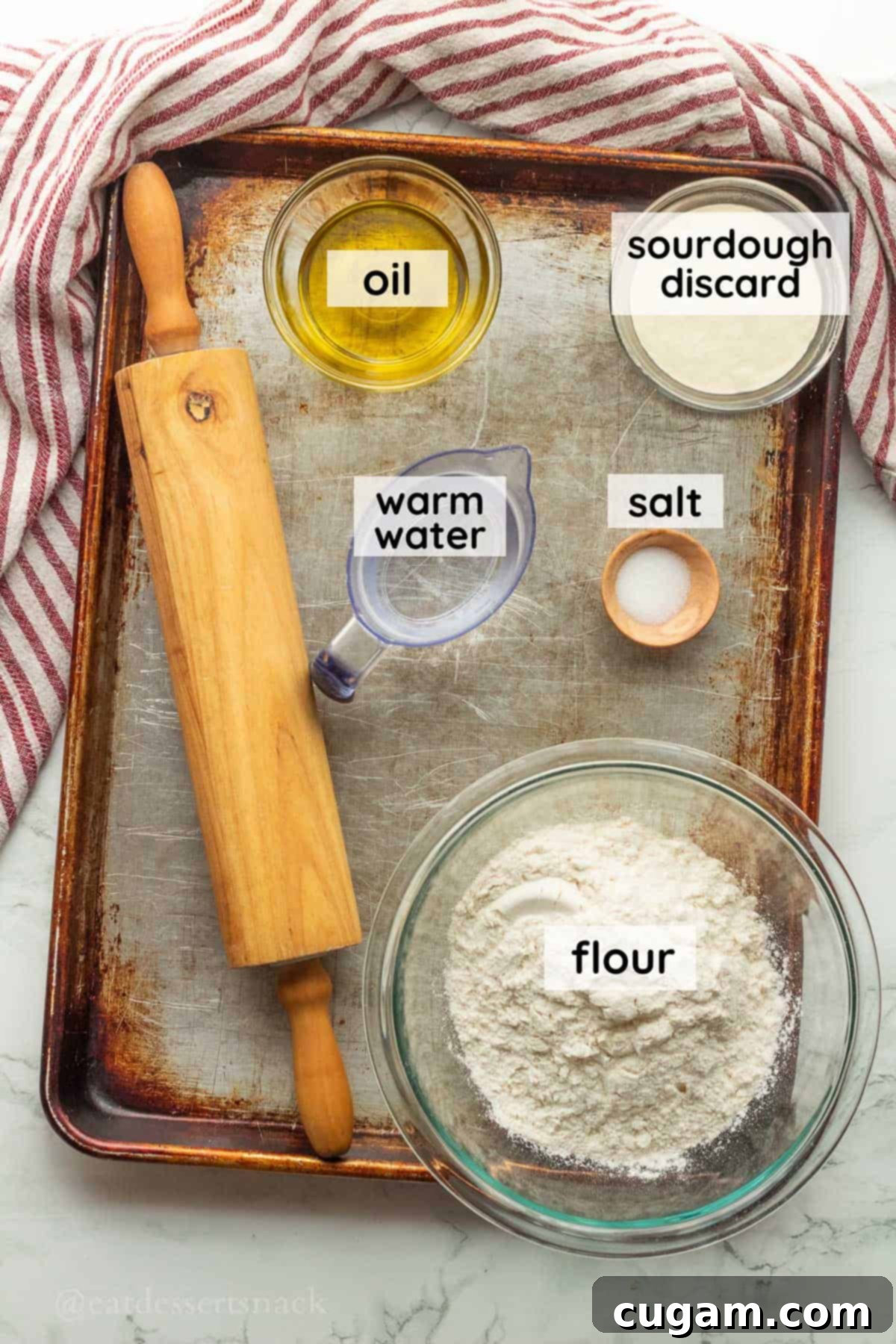 Essential ingredients for sourdough discard tortillas, including flour, sourdough discard, warm water, oil, and salt, arranged on a baking sheet.