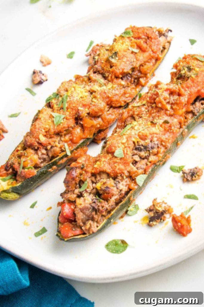 White plate with 2 stuffed zucchini boats. They are generously filled with a flavorful veggie and walnut mixture that remarkably resembles seasoned ground beef, lavishly covered with a vibrant tomato sauce, and garnished with fresh green herbs and a sprinkle of nutritional yeast. Bits and crumbs of the delicious filling are visible around the zucchini boats, hinting at the hearty texture.