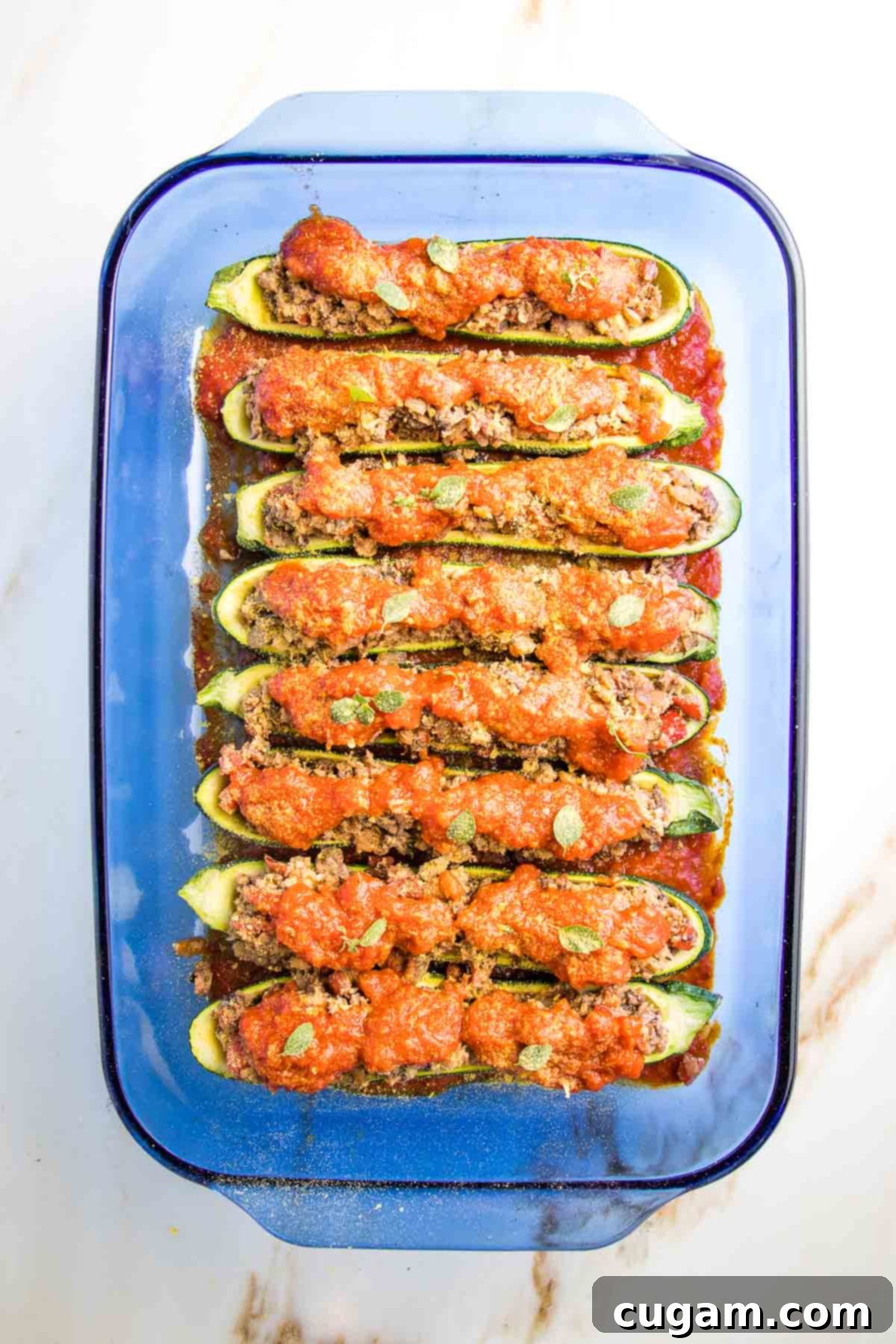 A glass baking dish showcasing stuffed zucchini boats, generously covered in marinara sauce and sprinkled with a golden layer of nutritional yeast, poised for baking.