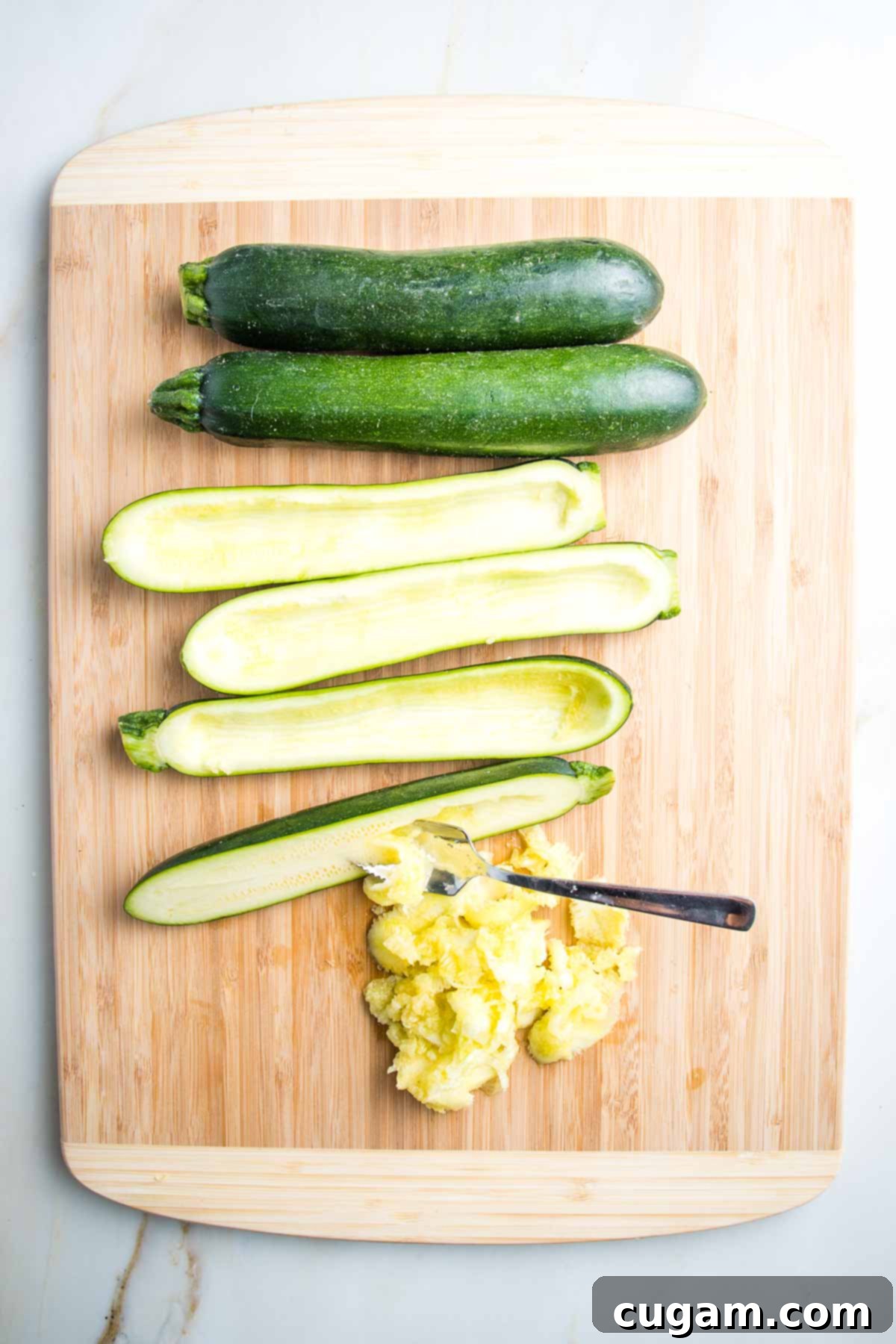 4 zucchinis on a cutting board. Two are whole, while two are cut in half lengthwise. The insides of one cut zucchini have already been scooped out, and a grapefruit spoon is actively scooping out the pulp from another, adding to a pile of removed pulp on the side.