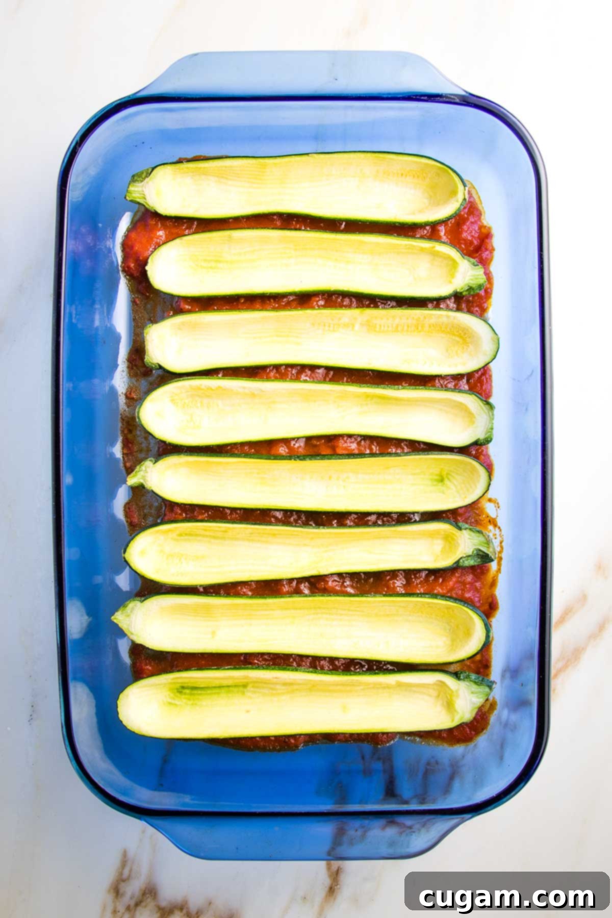 A 9 x 13 inch baking dish prepared with a thin, even layer of marinara sauce spread across the bottom, serving as a bed for the hollowed-out, empty zucchini boats which are neatly arranged on top of the sauce.