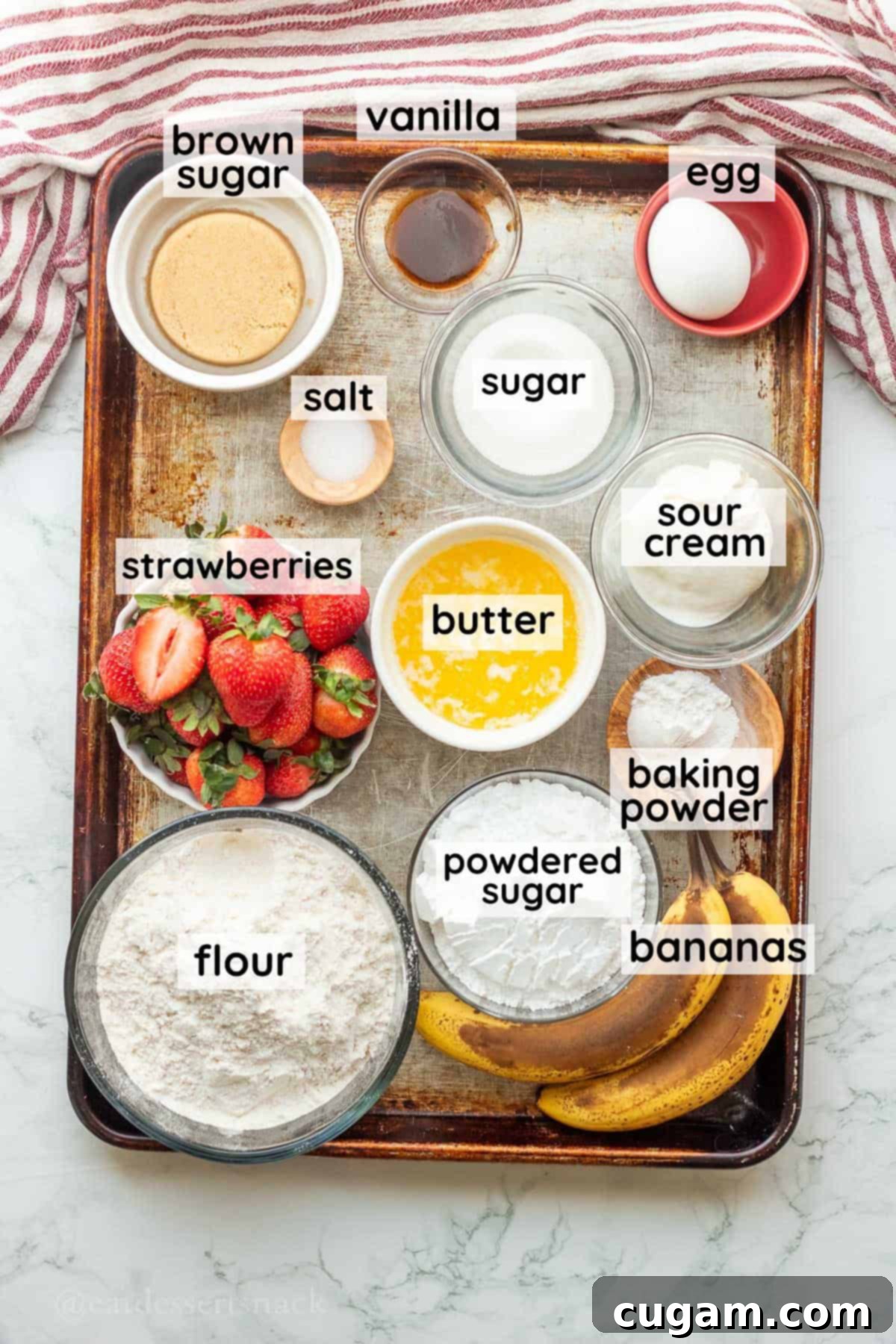 Labeled glass bowls containing the key ingredients for banana strawberry bread, including butter, strawberries, sour cream, and bananas, arranged on a metal baking pan.