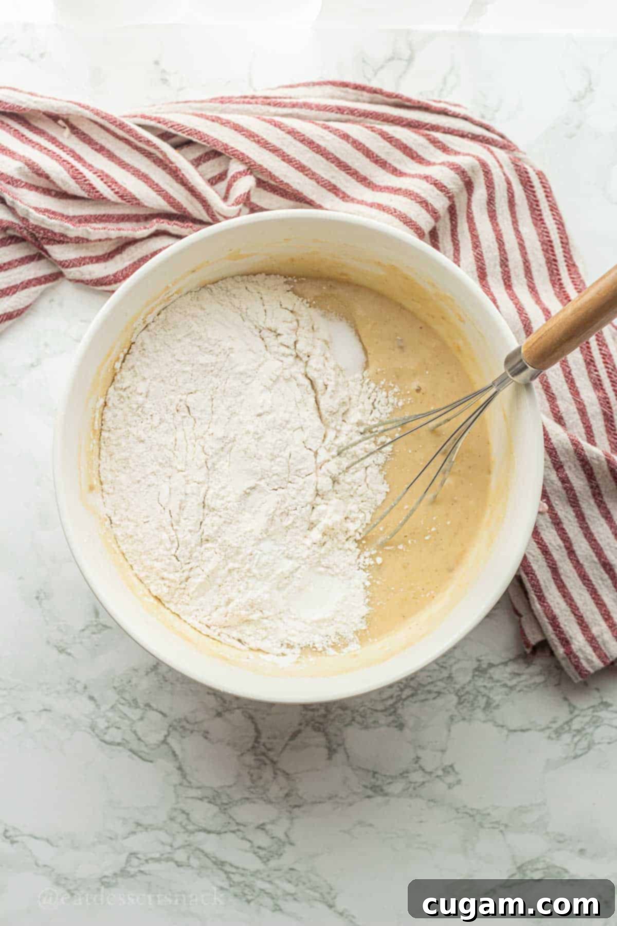 Flour, baking powder, and salt added on top of the wet ingredients in a white mixing bowl, ready to be combined for strawberry banana bread.