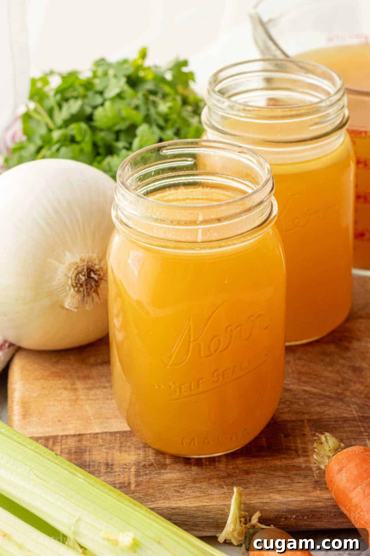 Chicken bone broth in jar on wood cutting board with fresh vegetables like carrots, celery, and onion.