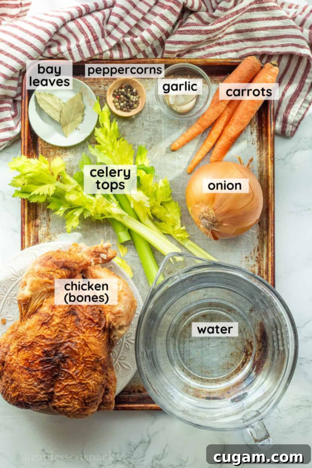 Labeled ingredients for chicken bone broth on a metal pan, including chicken bones, carrots, celery, onion, garlic, bay leaves, and peppercorns.