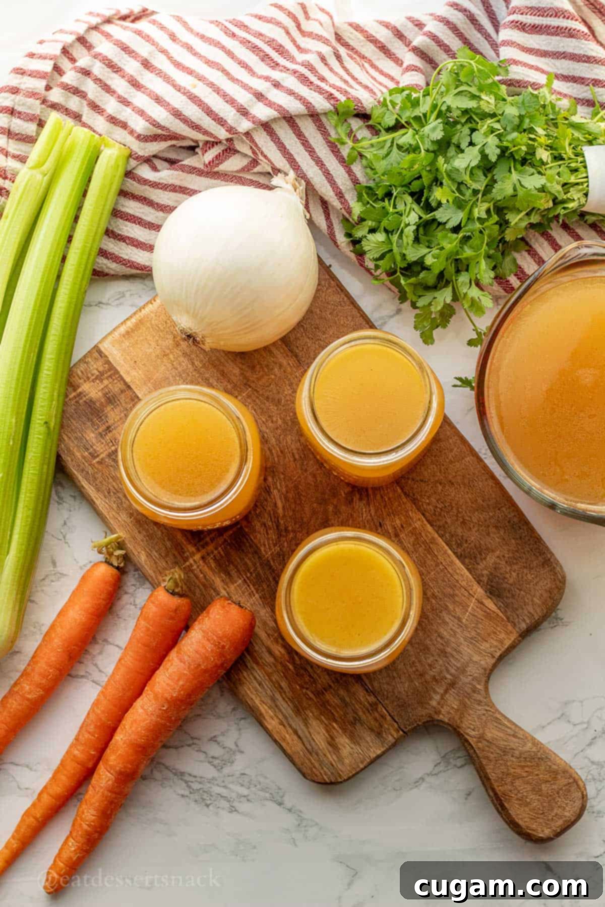 Jars of golden chicken bone broth on a wood cutting board, with fresh onion and carrots in the background.