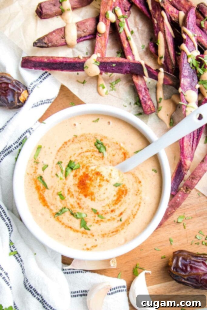 small white bowl with chipotle salad dressing, swirled with extra chipotle powder and chopped cilantro, surrounded by purple fries, dates, garlic, and a white dish towel with thin blue lines. Fries are drizzled with the creamy orange dressing.