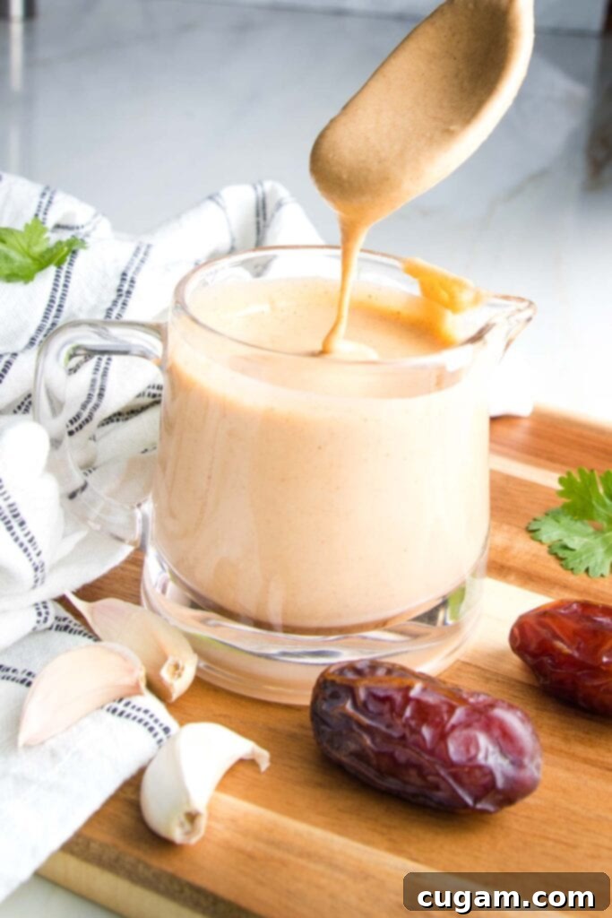 Glass pitcher filled with creamy light orange dressing. There is a spoon dripping dressing back into container...a few dates and cloves of garlic on the wooden board, dish towel or countertop surrounding the salad dressing.