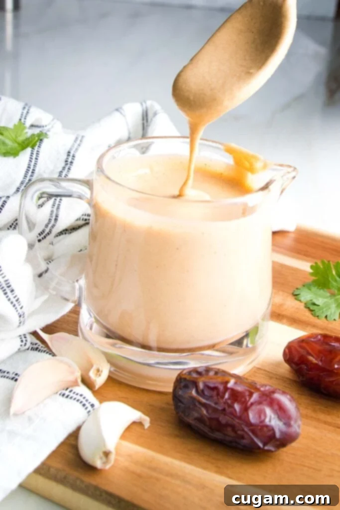 Glass pitcher filled with creamy light orange dressing.  There is a spoon dripping dressing back into container...a few dates and cloves of garlic on the wooden board, dish towel or countertop surrounding the salad dressing. 