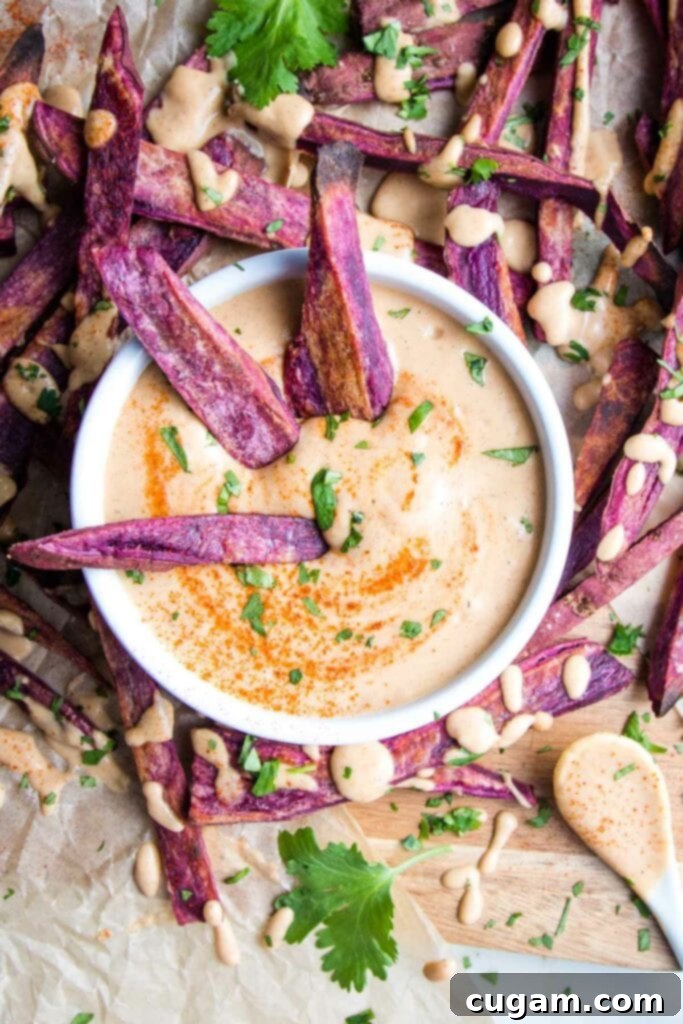 Purple sweet potato fries surrounding a small white bowl of chipotle mayo sauce that's also drizzled onto the fries.  A few fries are dipping into the sauce that's sprinkled with additional chipotle powder and chopped fresh cilantro. 
