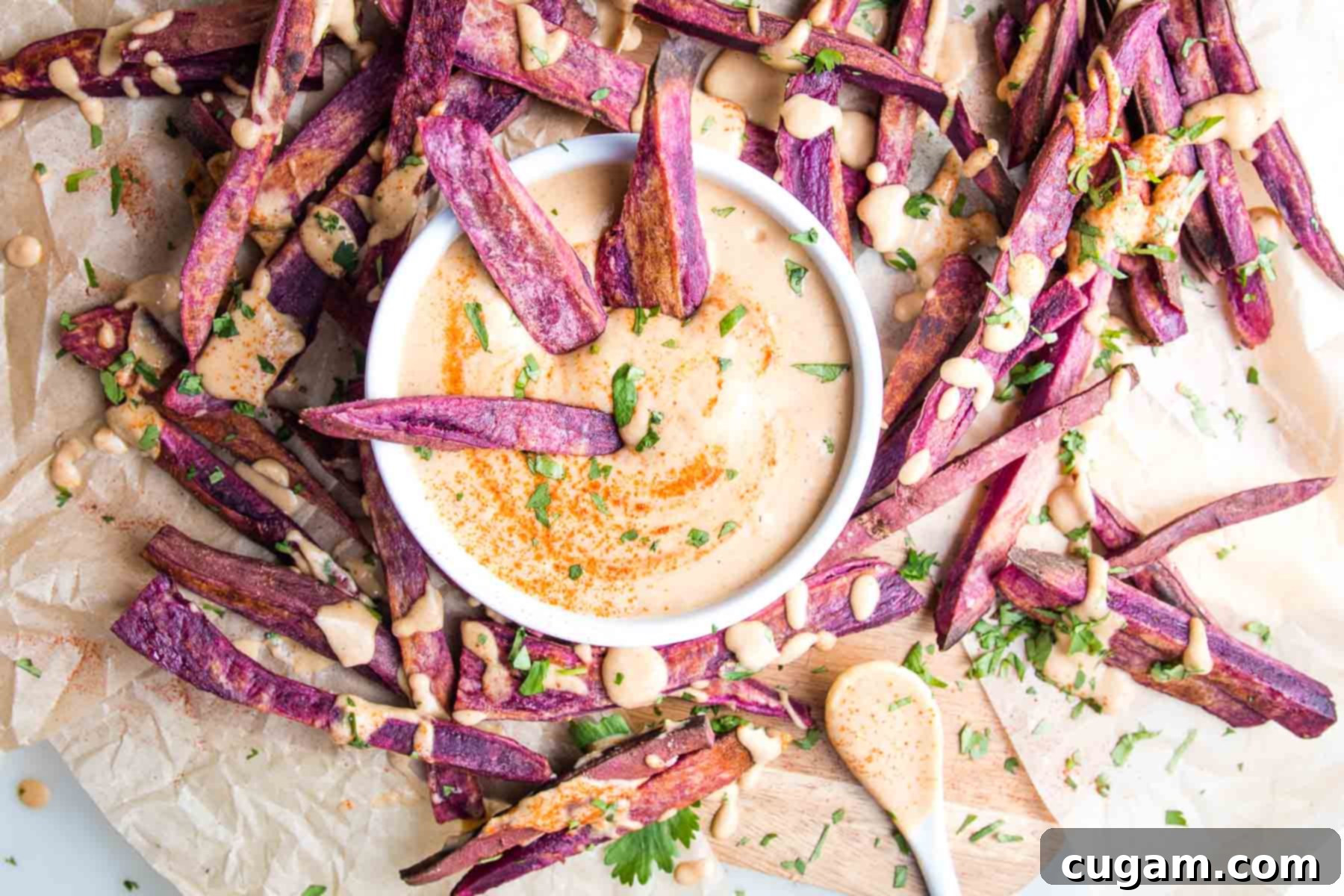 Purple sweet potato fries on unbleached parchment with a dish of chipotle dipping sauce and same orange sauce is drizzled over the fries.