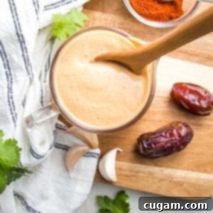 Small glass pitcher filled with orange creamy salad dressing, there are a couple of dates, a few cloves of garlic, some sprigs of cilantro and a small dish of chipotle powder surrounding the dressing that's on top of a wooden cutting board.