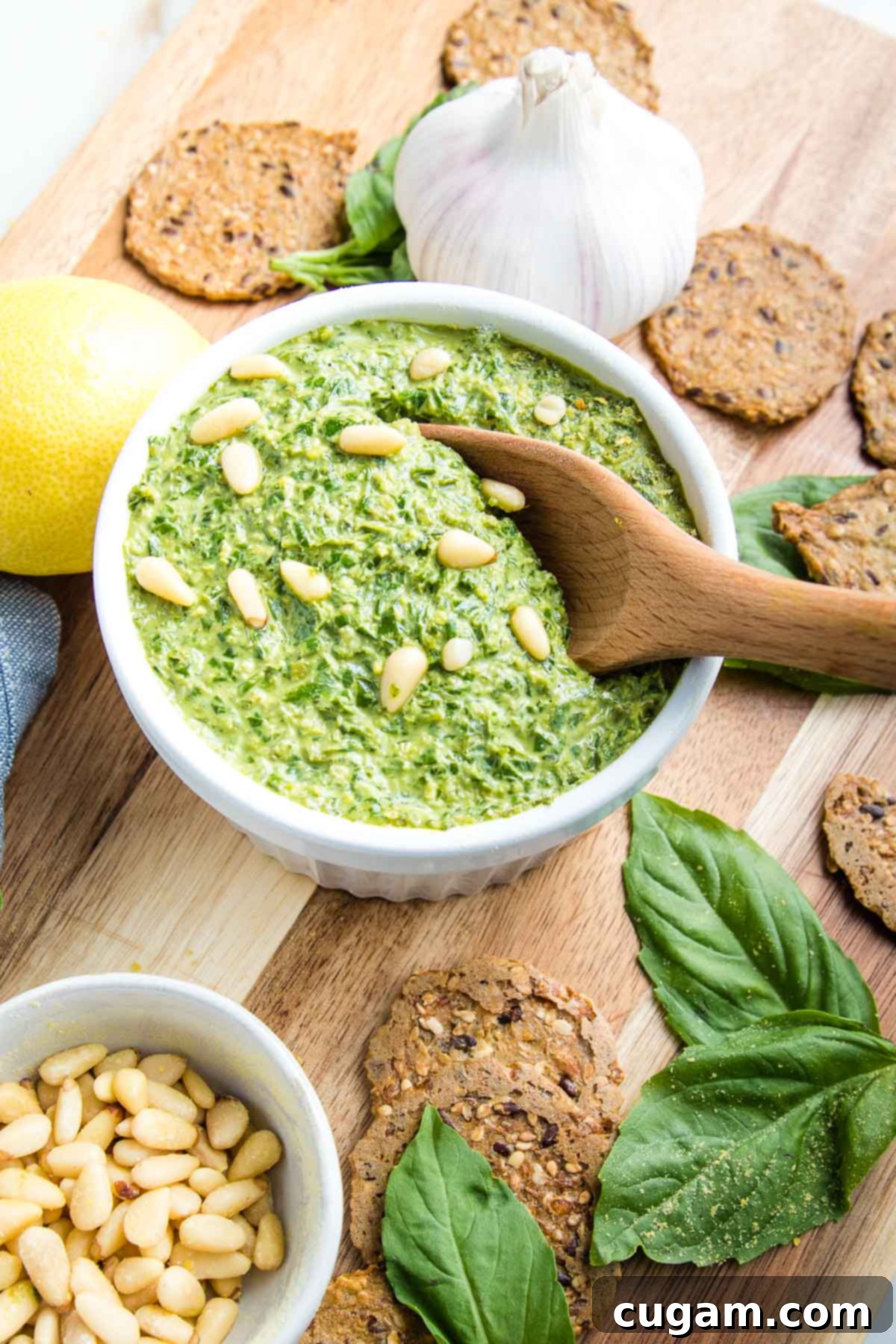 Small white bowl filled with bright green vegan basil pesto. Crackers, fresh basil leaves, a lemon, a head of garlic, and pine nuts are artfully arranged around the bowl on a rustic wooden board, creating an inviting food photography scene.