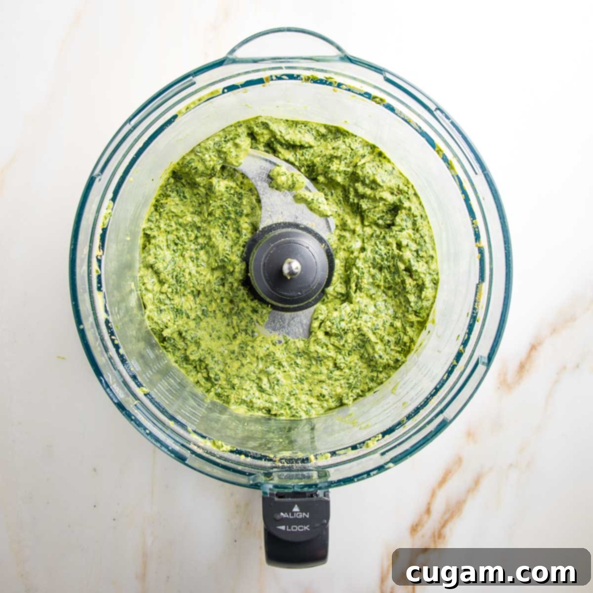 The food processor is running, blending the pesto to a creamy consistency as olive oil is slowly drizzled in, creating a smooth, emulsified green sauce.