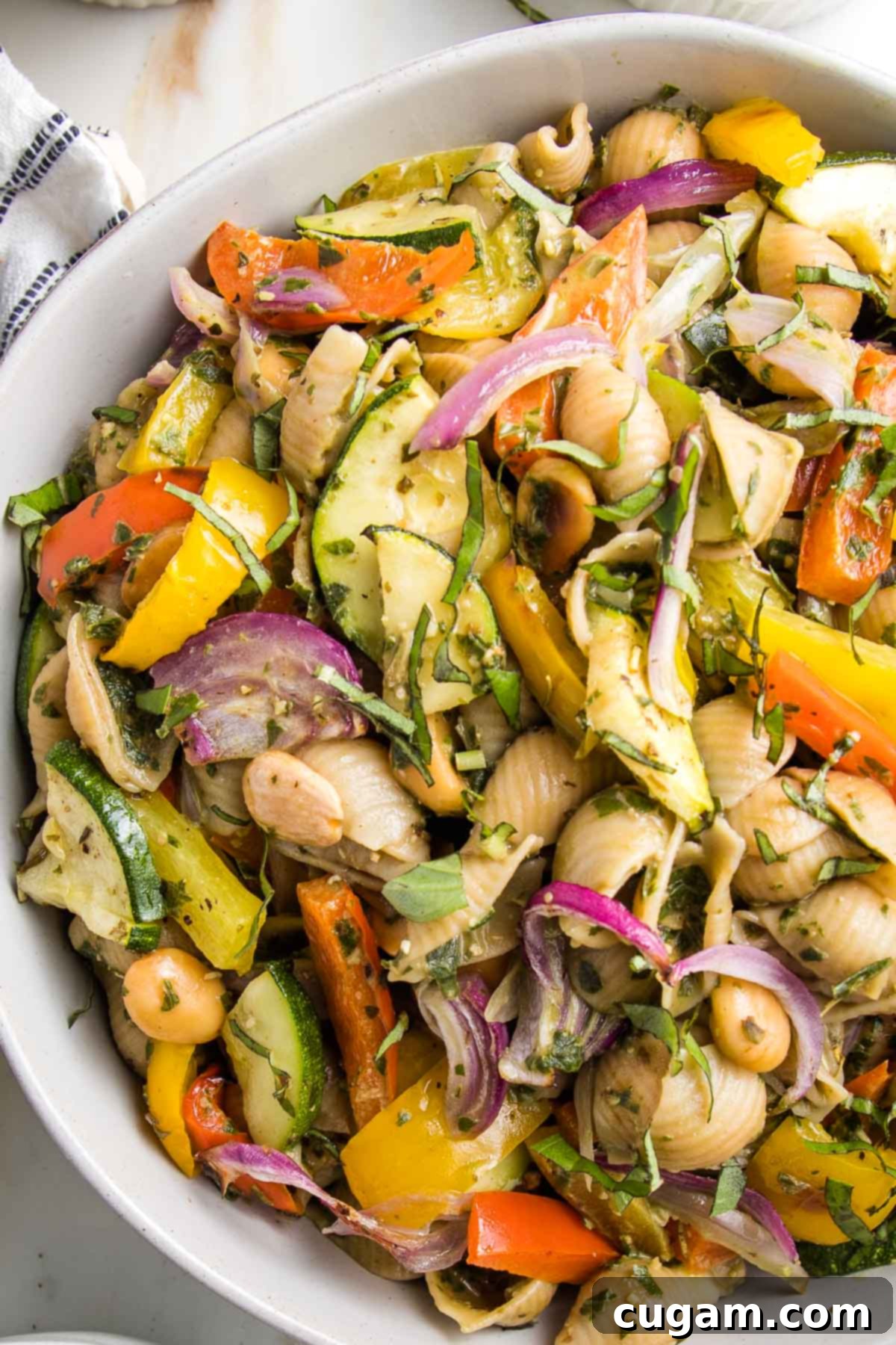 Close up of a bowl of pesto pasta salad with roasted vegetables.  It's colorful....red and yellow peppers, purple onions, green zucchini. The pasta is shells, the sauce is green.  All in a white bowl. 