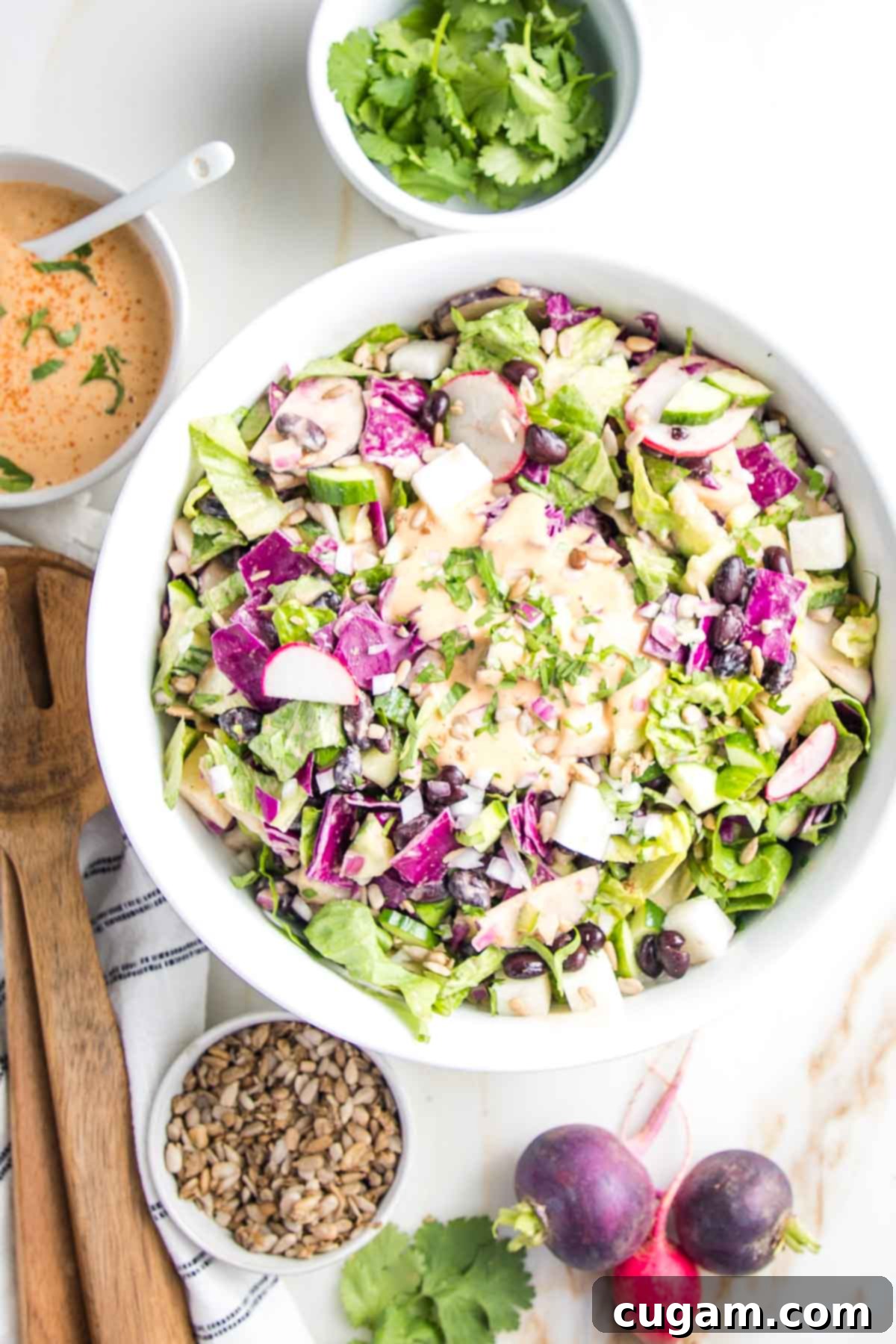 White bowl filled with colorful chopped salad and orange creamy chipotle dressing. A small bowl of fresh cilantro, one with additional chipotle dressing, smaller bowl with toasted sunflower seeds and some wooden salad servers surround the bowl of chopped salad, along with three fresh whole radishes and some sprigs of cilantro.