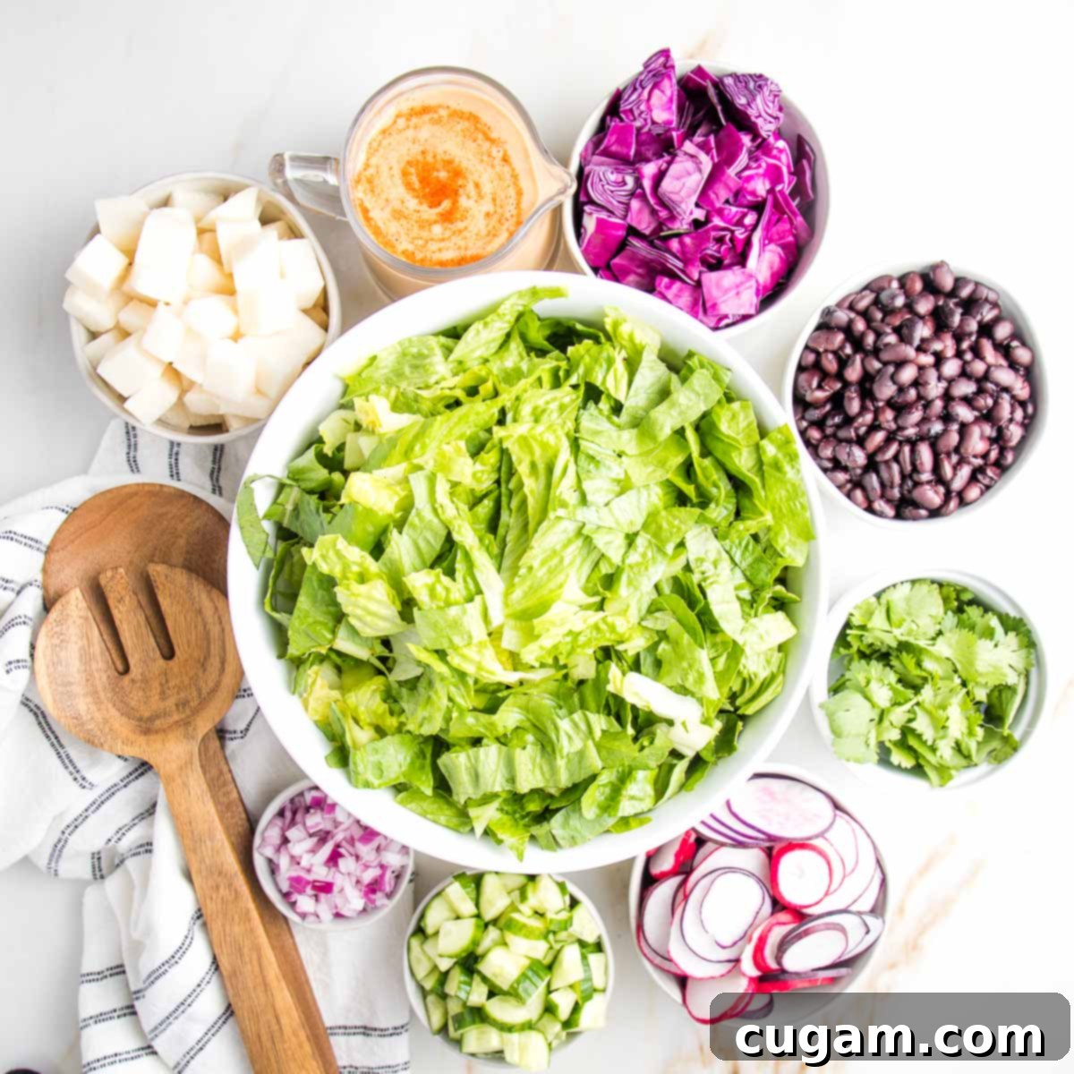 Large white bowl filled with chopped romaine in the center with small bowls of other ingredients all around: cubes of jicama, creamy chipotle dressing, chopped cabbage, black beans, fresh cilantro, sliced radishes, purple onions and a wooden set of salad tossers.