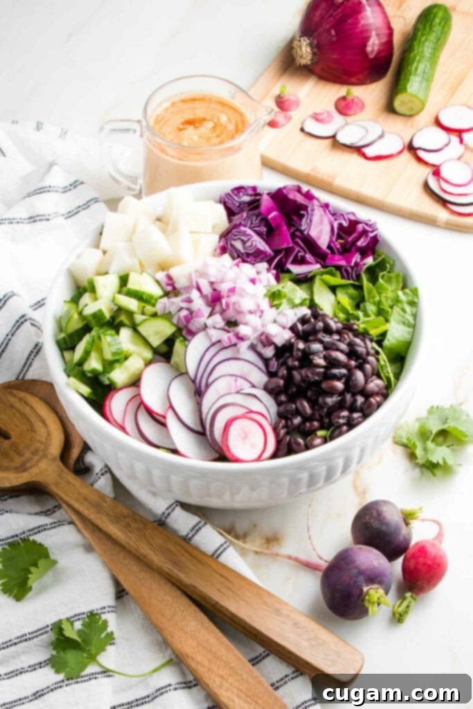 Large white bowl with wooden salad tossers on a white counter with a kitchen towel on the side that is white with blue stripes. The bowl is filled with sections of veggies: sliced radish, diced cucumbers, chopped jicama, red cabbage, fresh cilantro and black beans. THere is a cutting board in the background with slices of veggies and a small glass pitcher with chipotle dressing.