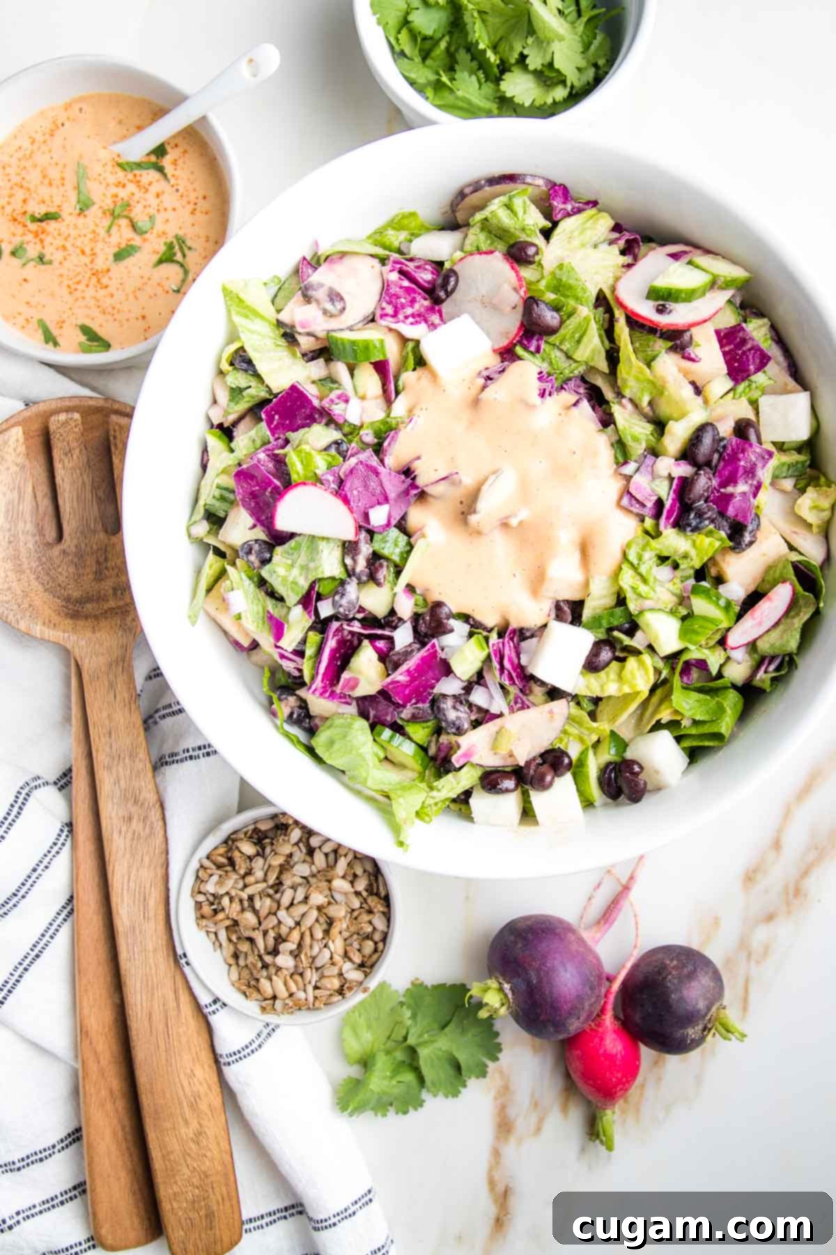 large chopped salad bowl with orange creamy dressing in the center. whole radish, fresh cilantro, chipotle dressing, sunflower seeds and wooden salad servers surround the colorful bowl.