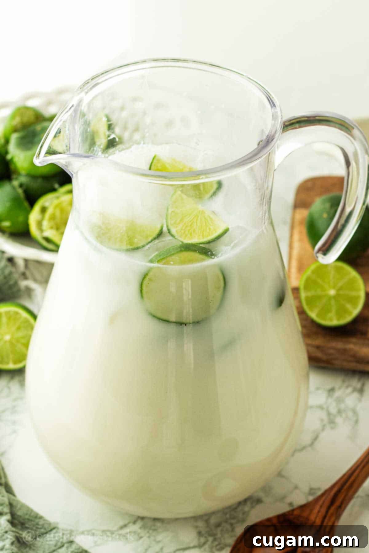 A large pitcher of creamy Brazilian limeade with lime wedges on a marble countertop, ready to be served.