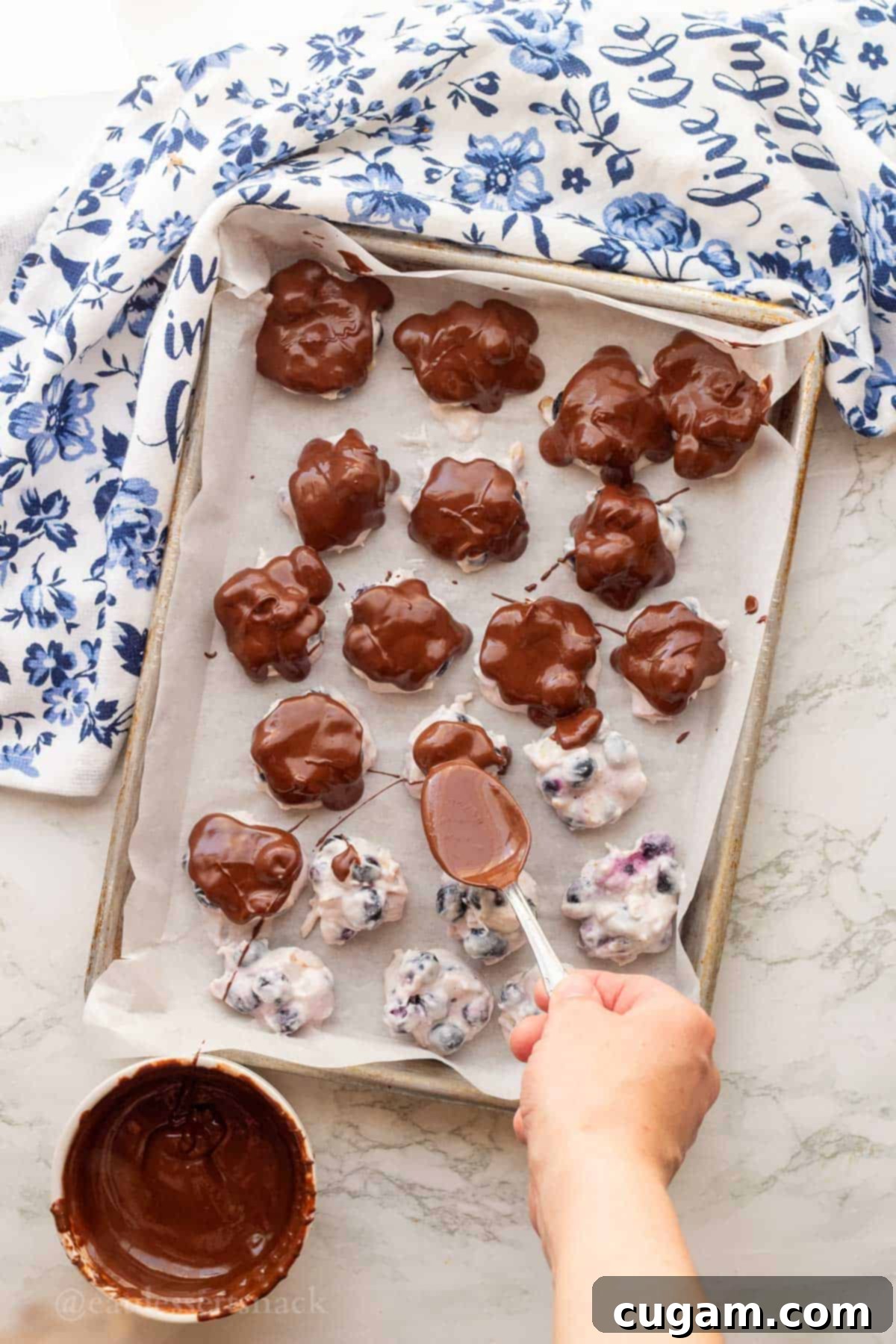 Spooning melted chocolate over frozen blueberry yogurt clusters.