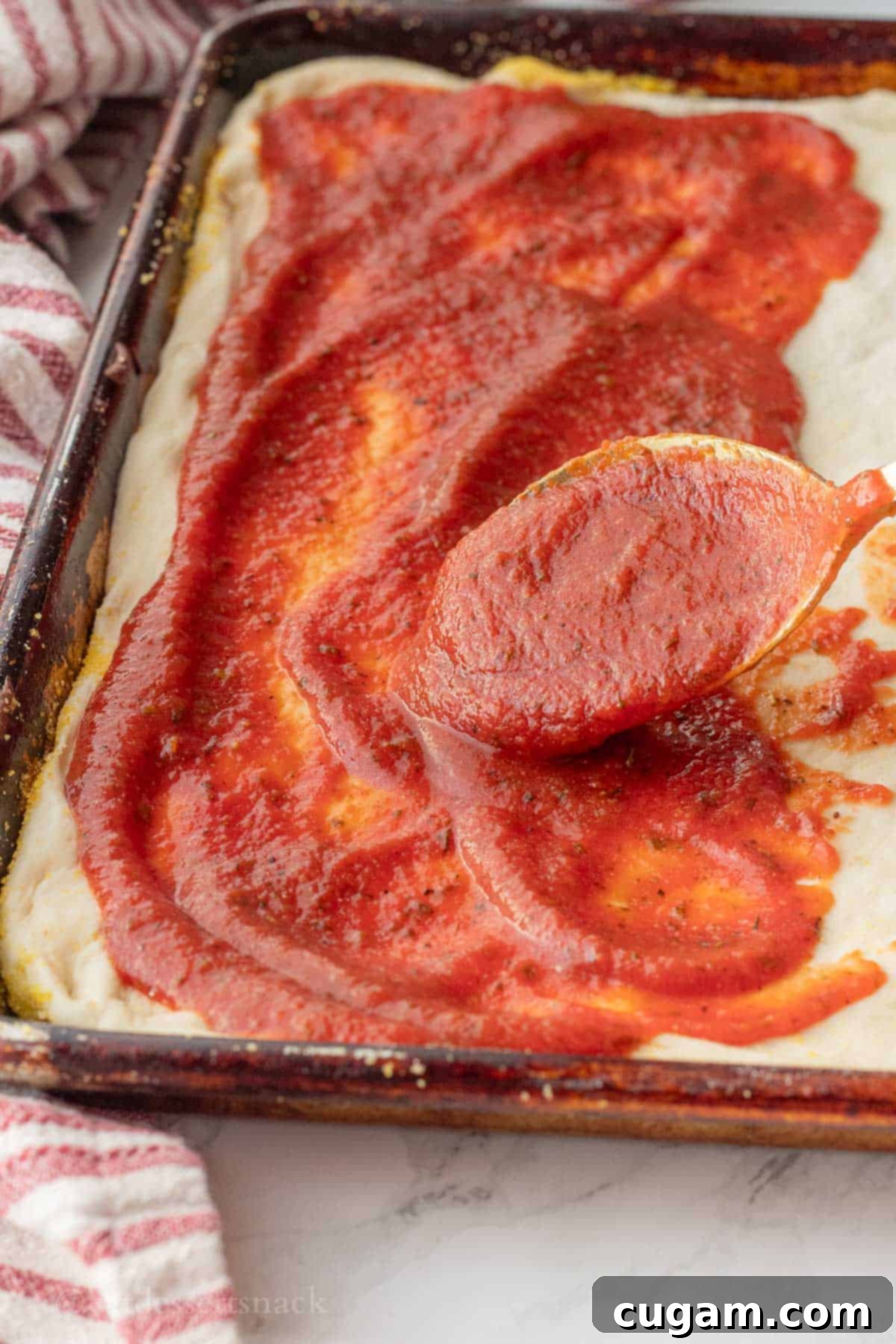 A spoon spreading rich, red tomato paste pizza sauce evenly over a raw pizza dough, ready for toppings.
