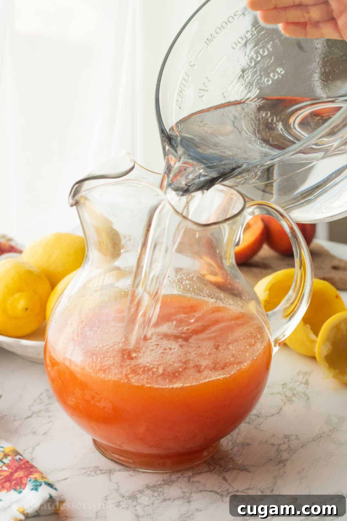Clear water being poured into a glass pitcher already containing a mix of peach juice and other lemonade ingredients.