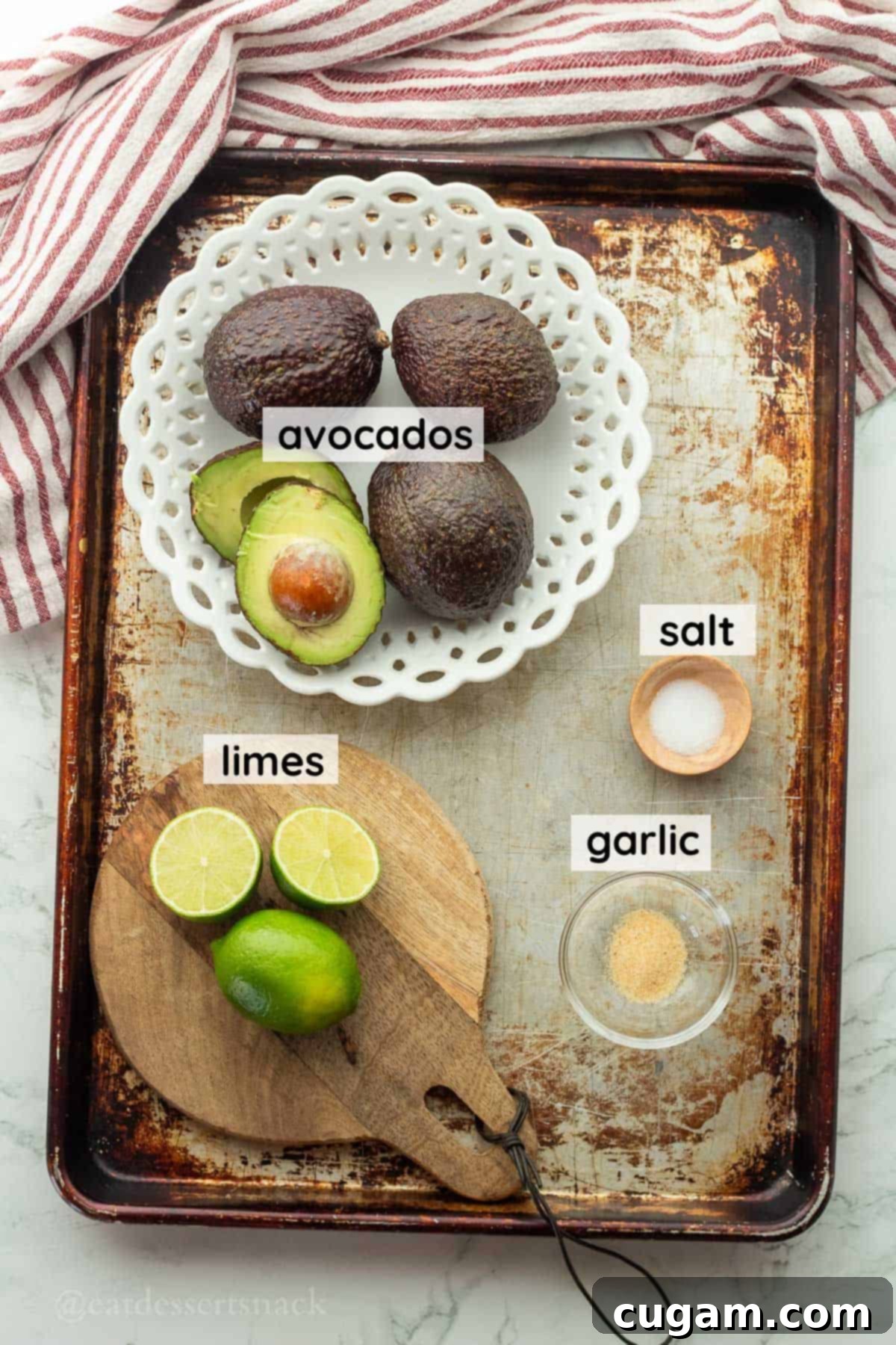 Labeled ingredients for guacamole, including ripe avocados, fresh lime, garlic powder, and salt, arranged on a metal pan.