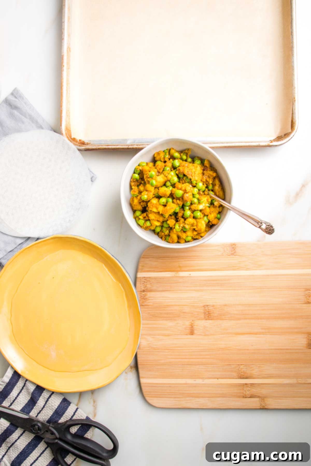 Everything ready to begin making samosas: rimmed baking tray lined with parchment, stack of rice paper wrappers, rimmed dish filled with water, samosa filling, small wooden board, kitchen scissors and a dish towel.