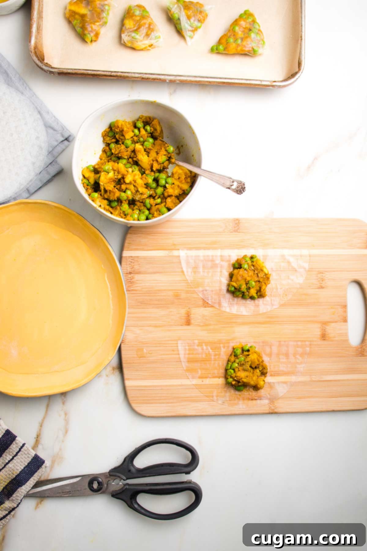 Rimmed baking sheet lined with parchment, a bowl of potato samosa filling, a yellow dish filled with water, a pair of kitchen scissors, and a wooden board with two half moons of rice paper each with a scoop of filling.